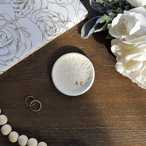 May include: White ceramic ring dish with an embossed floral design and the initials "AT" in gold lettering. The dish is on a dark brown wooden surface.