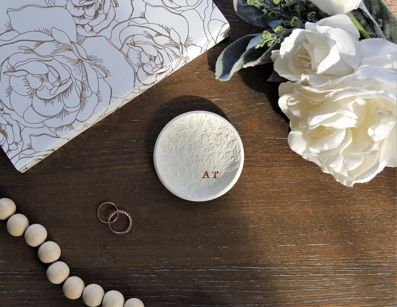 May include: White ceramic ring dish with an embossed floral design and the initials "AT" in gold lettering. The dish is on a dark brown wooden surface.
