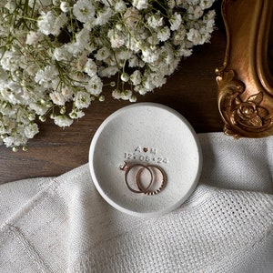 May include: A white ceramic ring dish with the initials "A & M" and the date "12.05.24" inscribed. Two gold rings are resting inside the dish.