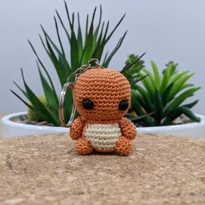 May include: A crocheted orange and white character keychain with black eyes. The keychain is hanging from a silver key ring.