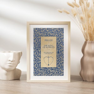May include: A framed print of a book cover with a blue background and gold leaf design. The title of the book is "The Song of Achilles" by Madeline Miller. The quote "He is half of my soul, as the poets say" is printed below the title.