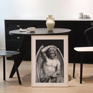 May include: A black and white framed print of a sculpture of a winged figure with a sorrowful expression. The sculpture is set against a dark background.