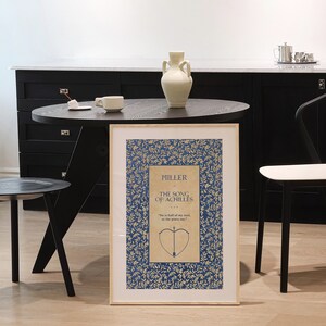 May include: A framed print of the book cover for "The Song of Achilles" by Madeline Miller. The cover features a blue background with a gold leaf pattern and a quote from the book. The title of the book is in gold lettering.