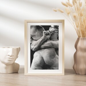 May include: A framed black and white print of a classical sculpture depicting two figures in a dynamic embrace. The artwork is set in a light wood frame, displayed on a wooden surface, and is accompanied by a decorative vase.