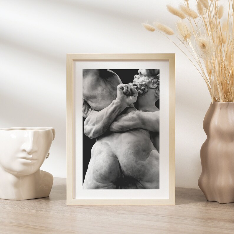 May include: A framed black and white print of a classical sculpture depicting two figures in a dynamic embrace. The artwork is set in a light wood frame, displayed on a wooden surface, and is accompanied by a decorative vase.