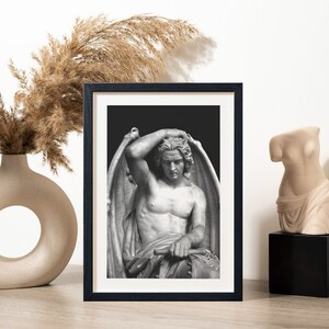 May include: Black and white framed print of a sculpture of a winged figure with a sorrowful expression. The figure is seated and has its hand on its head.