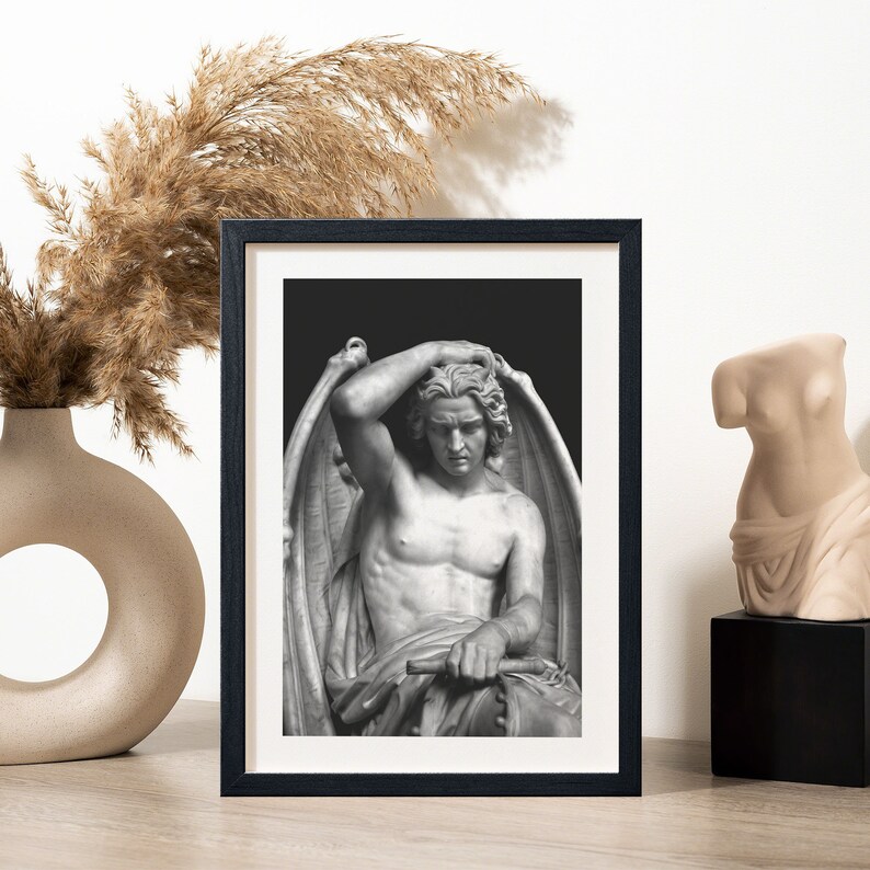 May include: Black and white framed print of a sculpture of a winged figure with a sorrowful expression. The figure is seated and has its hand on its head.