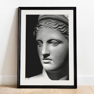 May include: Black framed print featuring a grayscale image of a classical sculpture bust. The sculpture depicts a woman's face with detailed features, including a serene expression and wavy hair. The artwork is set against a dark background.