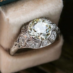 May include: Close-up of a vintage engagement ring with a large, round-cut diamond as the centerpiece. The ring features intricate filigree and small accent diamonds set in a white metal band, likely platinum or white gold. The ring is displayed in a velvet ring box.