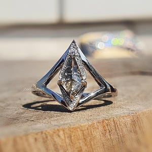 May include: A silver-toned ring featuring a central kite-shaped stone set within a geometric, diamond-shaped frame. The frame is accented with small, clear stones along the edges, creating a sharp, angular silhouette on a polished metal band.
