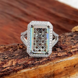 Puede incluir: Un anillo de plata con una gran piedra preciosa transparente de corte esmeralda rodeada de diamantes más pequeños. El anillo está colocado sobre una superficie de madera.