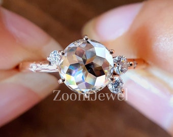 May include: A rose gold ring featuring a large, round, faceted center stone with a cluster of three smaller, clear, round-cut stones on one side. The band is thin and metallic, and the text ZoomJewel is visible in white at the bottom center.