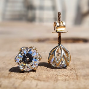 May include: A pair of gold stud earrings featuring round, faceted clear stones. One earring is positioned face-up, displaying the brilliant cut, while the other stands upright, revealing the six-prong basket setting and the threaded post back.