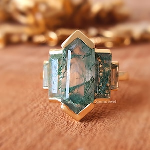 May include: A gold ring with a green moss agate gemstone. The ring has a geometric design with a pointed top and a rectangular base.