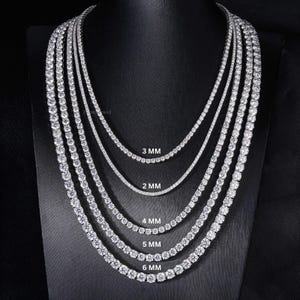 May include: A display of several diamond tennis necklaces, each with varying stone sizes. The necklaces are arranged in a cascading fashion, showcasing the different millimeter sizes: 2 MM, 3 MM, 4 MM, 5 MM, and 6 MM. The jewelry is displayed on a black stand.