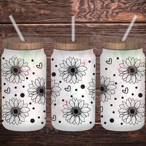 Puede incluir: Tres vasos de vidrio con tapas de madera y pajitas. Cada vaso presenta un fondo blanco con ilustraciones de girasoles negros, corazones y lunares. Los vasos están dispuestos sobre una superficie de madera.