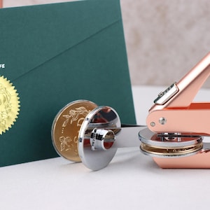 Puede incluir: Un sello de cera en oro rosa con un mango a juego, junto a un sobre verde con un sello de lámina dorada. El sello tiene el texto "S & D" y las palabras "THE SIMPLE LIFE".