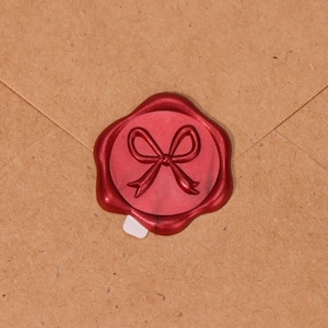 May include: A red wax seal featuring a bow design, applied to a brown envelope. The seal is circular with a raised bow image in the centre. The wax has a glossy finish, and the envelope is made of textured paper.