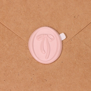 May include: A pale pink wax seal with a bow design, set against a brown paper envelope. The oval seal features a detailed bow and ribbon design, ideal for adding a decorative touch to letters and crafts.