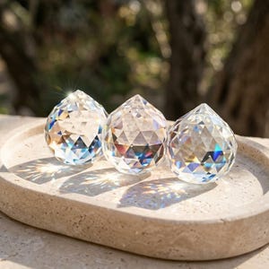 Könnte beinhalten: Drei klare, facettierte Kristallkugeln mit spitzer Oberseite, die in einer Reihe angeordnet sind. Jede Kugel hat eine geometrische, mehrfach facettierte Oberfläche, die das Licht bricht und helle, prismatische Reflexionen erzeugt.