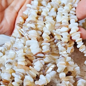 Könnte beinhalten: Nahaufnahme einer Kette aus Muschelsplitterperlen. Die Perlen sind eine Mischung aus Weiß, Creme und Braun und haben eine natürliche, unregelmäßige Form. Die Perlen sind aufgefädelt und erzeugen einen strukturierten und organischen Look.