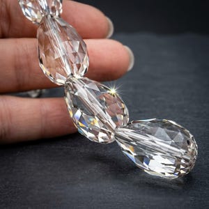 Könnte beinhalten: Eine Kette aus klaren, facettierten Kristallperlen in Tropfenform. Jede Perle weist mehrere geometrische Schliffe auf, die das Licht reflektieren und für ein brillantes Funkeln sorgen. Die Perlen sind in einer Reihe angeordnet und zeigen ihre Transparenz.
