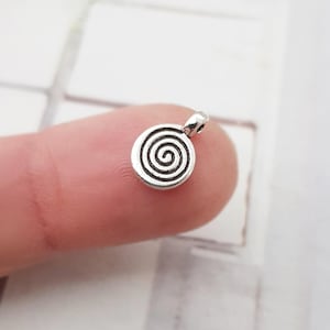 Könnte beinhalten: Silberner Anhänger mit Spiralmuster. Der Anhänger ist rund und hat oben eine Öse zum Anbringen an einer Halskette oder einem Armband.