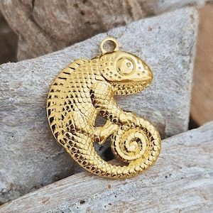 Könnte beinhalten: Goldfarbener Chamäleon-Anhänger mit detaillierten Schuppen. Der Anhänger hat einen eingerollten Schwanz und eine Schlaufe zum Aufhängen. Das Chamäleon liegt auf einem Stück verwittertem Holz.