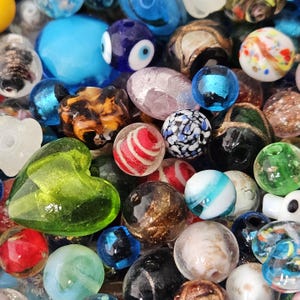 Könnte beinhalten: Eine Sammlung verschiedener Glasperlen in unterschiedlichen Formen, Größen und Farben. Das Sortiment umfasst eine markante grüne herzförmige Perle, eine blaue Augenperle, klare Perlen mit Goldpartikeln sowie kugelförmige Perlen mit roten, weißen und blauen Wirbeln.