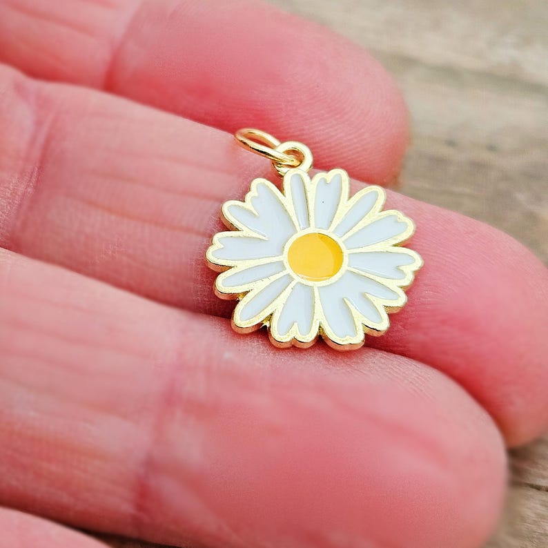 May include: A gold-colored daisy charm with white petals and a yellow center. The charm has a small loop at the top for attaching to a necklace or bracelet. The daisy is a classic floral design.