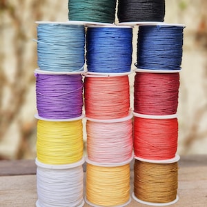 Könnte beinhalten: Ein Stapel bunter Kordelspulen in verschiedenen Farbtönen, darunter Blau, Lila, Rot, Gelb und Grün. Die Kordel ist eng auf weißen Kunststoffspulen aufgewickelt, was eine ansprechende Darstellung ergibt. Der Hintergrund ist unscharf, wodurch die lebendigen Farben der Kordel hervorgehoben werden.