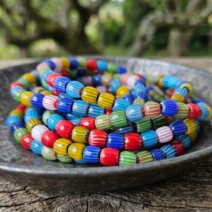 Könnte beinhalten: Ein gewickeltes Armband aus kleinen, gerippten, mehrfarbigen Glasperlen. Die Perlen weisen vertikale Rillen auf und sind in einem sich wiederholenden Muster aus leuchtendem Rot, Gelb, Blau, Grün, Rosa und Weiß angeordnet, was für eine lebendige Textur sorgt.