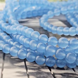 Könnte beinhalten: Ein Strang aus glatten, runden, hellblauen Perlen, die in einer spiralförmigen Anordnung liegen. Die Perlen haben ein halbtransparentes Aussehen mit einer glänzenden Oberfläche, die das Licht reflektiert. Der Strang ist lang und flexibel.