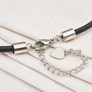 Könnte beinhalten: Eine Halskette aus schwarzem Lederband mit silberfarbenen Metallelementen, bestehend aus zwei zylindrischen Endkappen, einem Karabinerverschluss, einer kurzen Verlängerungskette und einem kleinen, glatten, herzförmigen Anhänger am Kettenende.