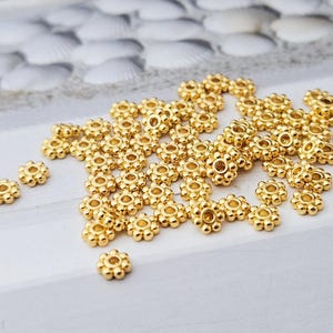 Könnte beinhalten: Eine Ansammlung kleiner, goldfarbener, blütenförmiger Perlen. Jede Perle hat ein zentrales Loch und ist von abgerundeten Blütenblättern umgeben. Die Perlen sind auf einer weißen Oberfläche verstreut und erzeugen ein zartes und dekoratives Aussehen. Ideal für Schmuckherstellung und Basteln.