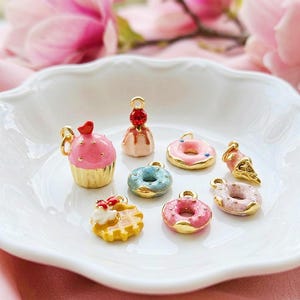 Könnte beinhalten: Eine Sammlung kleiner Metallanhänger in Form von Desserts, darunter ein rosa Cupcake, eine Waffel mit Schlagsahne, drei glasierte Donuts in Rosa und Blau, ein kleiner Kuchen mit einem roten Schmuckstein und eine Miniatur-Eistüte mit Goldakzenten.