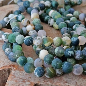 Könnte beinhalten: Eine Sammlung runder Perlen mit mattem Finish in verschiedenen Schattierungen von Waldgrün, Salbei, Creme und Weiß. Die Perlen weisen natürliche, gesprenkelte Muster und Einschlüsse auf, die eine texturierte Optik auf den Oberflächen erzeugen.