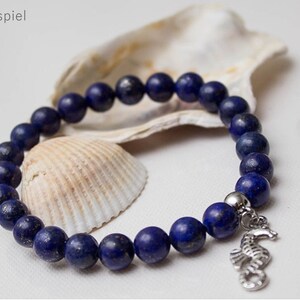 May include: A blue lapis lazuli beaded bracelet with a silver seahorse charm. The bracelet is displayed on a seashell.