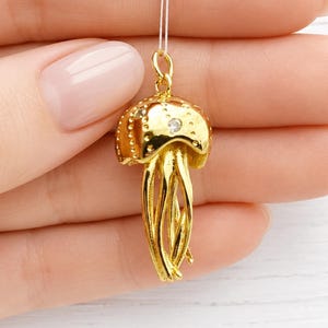 Könnte beinhalten: Goldfarbener Quallenanhänger mit kleinem Diamantakzent. Der Anhänger ist an einem dünnen, weißen Faden aufgehängt. Das Quallendesign hat eine gewölbte Oberseite mit kleinen, erhabenen Punkten und fließenden Tentakeln.