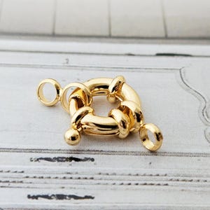Könnte beinhalten: Ein goldfarbener, quadratischer Schmuckverschluss mit abgerundeten Kanten und einem kreisförmigen Ring an jedem Ende. Der Verschluss hat eine polierte Oberfläche und ist für Halsketten oder Armbänder konzipiert.