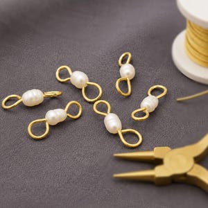 Könnte beinhalten: Fünf goldfarbene Schmuckverbinder aus Metall, jeweils mit einer einzelnen weißen, ovalen Perle in der Mitte. Die Verbinder haben an jedem Ende eine Öse zur Befestigung an Ketten oder anderen Schmuckkomponenten und liegen auf einem grauen Stoff.