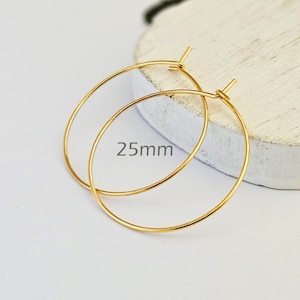 Könnte beinhalten: Ein Paar goldfarbene Creolen. Die Ohrringe sind kreisförmig und haben einen Durchmesser von 25 mm. Sie werden auf einer weißen Oberfläche präsentiert.