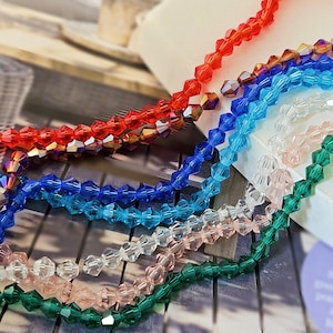 Könnte beinhalten: Stränge facettierter Perlen in verschiedenen Farben, darunter Rot, Blau, Klar, Rosa und Grün. Die Perlen sind aufgefädelt und erzeugen einen schimmernden Effekt. Ideal für die Schmuckherstellung oder Bastelprojekte.