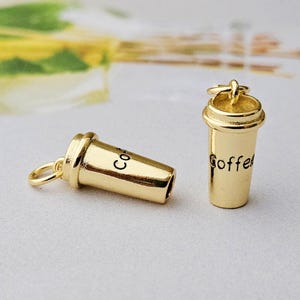 May include: Two gold-colored coffee cup charms. One charm is lying on its side, and the other is upright. The word "Coffee" is written on the upright charm, and the word "Co" is written on the other charm.