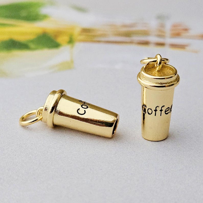 May include: Two gold-colored coffee cup charms. One charm is lying on its side, and the other is upright. The word "Coffee" is written on the upright charm, and the word "Co" is written on the other charm.