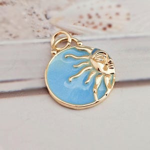 May include: A gold-colored pendant featuring a celestial design. The pendant has a light blue enamel background with a gold sun and moon face. The pendant is round and has a loop for attaching to a chain or bracelet.