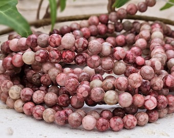 May include: Close-up of a pile of round, pink and burgundy gemstone beads. The beads have a mottled appearance, with varying shades of pink and burgundy. The beads are strung together, and the image is well-lit.