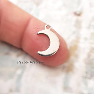 Könnte beinhalten: Ein kleiner, silberfarbener Halbmond-Anhänger mit einer Schlaufe zum Befestigen an Schmuck. Der Anhänger ist glatt und glänzend und reflektiert das Licht. Der Anhänger wird zwischen zwei Fingern gehalten.