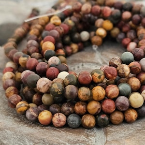 Könnte beinhalten: Nahaufnahme eines Strangs runder Natursteinperlen in verschiedenen Erdtönen. Die Perlen sind eine Mischung aus Rot, Braun, Gelb und Grün mit mattem Finish. Die Perlen haben einen Durchmesser von etwa 0,8 cm.