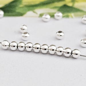 Könnte beinhalten: Nahaufnahme von glänzenden, silberfarbenen Perlen, die auf einem dünnen Draht aufgereiht sind. Die kugelförmigen Perlen haben ein kleines Loch. Mehrere lose Perlen sind im Hintergrund verstreut. Die Perlen sind wahrscheinlich für die Schmuckherstellung.